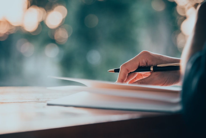 a woman writes in a notebook on a small wooden table in front of a blurred window with lights in the background 