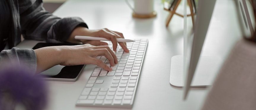 Erudus users can connect and communicate via our New Slack Channel women typing on a mac wireless keyboard with an apple pen in hand and an ipad infront of the keyboard