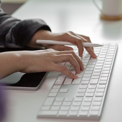 women typing on a mac wireless keyboard with an apple pen in hand and an ipad infront of the keyboard 