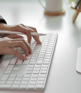 Erudus users can connect and communicate via our New Slack Channel  women typing on a mac wireless keyboard with an apple pen in hand and an ipad infront of the keyboard