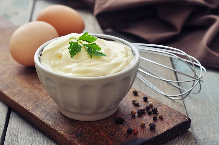 thin dark wood display board layed on a wooden table with two eggs resting on it next to a pot of creamy mayonnaise and sprinkled with peppercorns 
