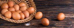 open wicker basket filled with eggs spilling out onto a dark wooden oak table and a small white feather 