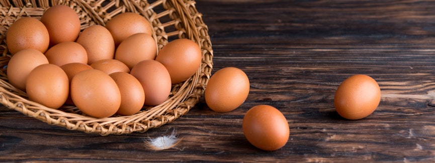 open wicker basket filled with eggs spilling out onto a dark wooden oak table and a small white feather 