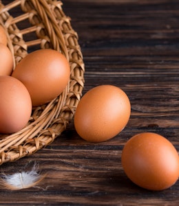 open wicker basket filled with eggs spilling out onto a dark wooden oak table and a small white feather 