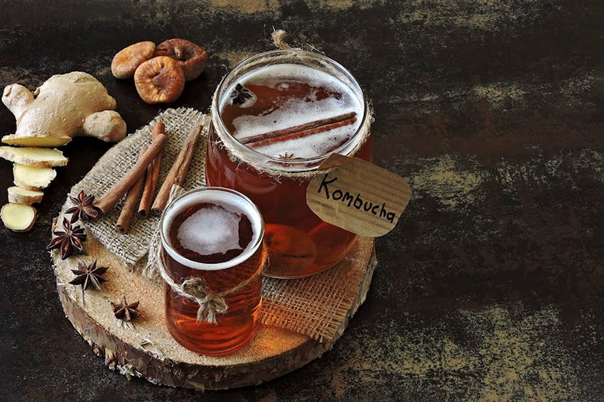 large jar of ginger spiced kombucha next to a smaller glass of kombucha next to 4 sticks of ginger and a diced ginger root on top of a circular platform on a dark rustic table 