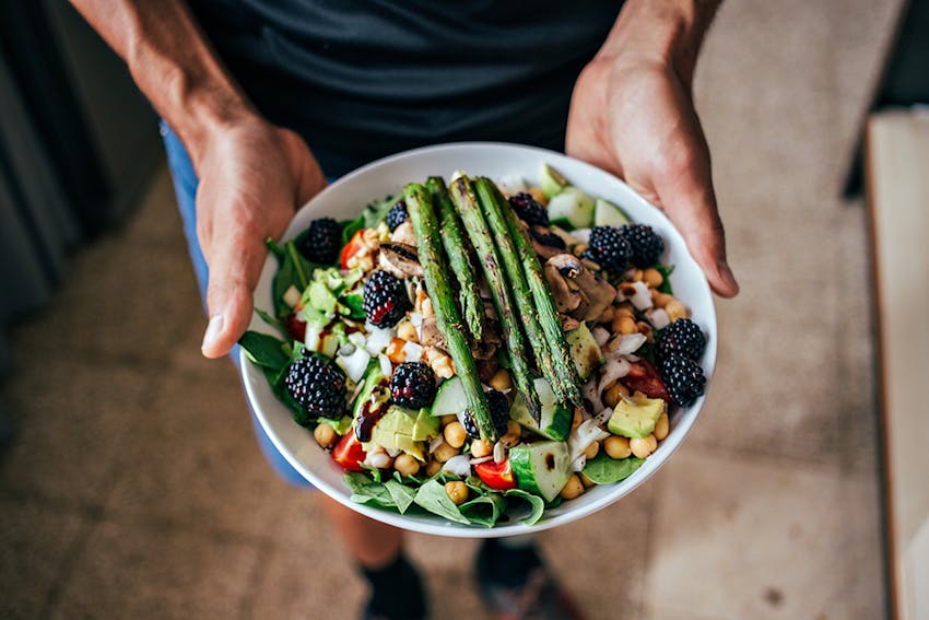 man holding a plate containing a varied and colourful salad topped with asparagus and blackberries 