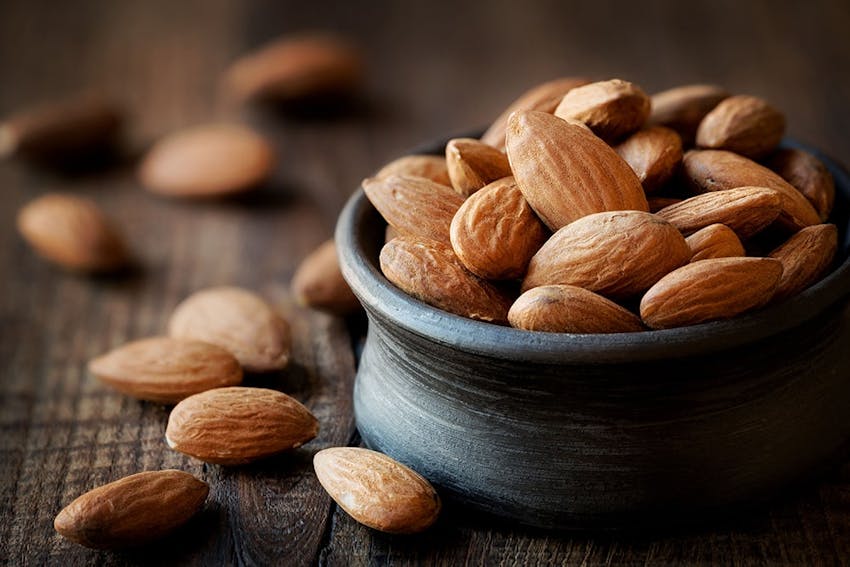 Small black rounded pot of almonds with almonds scattered around the pot on a wooden table 