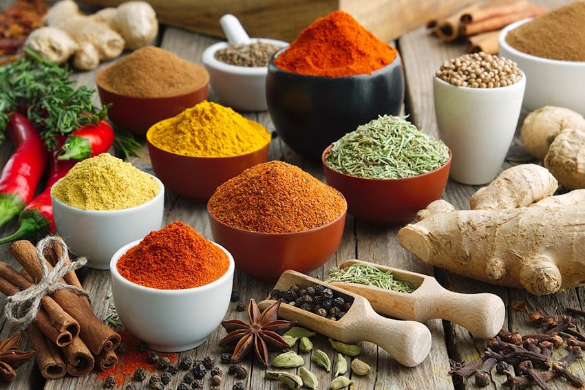 wooden table with an array of different sized pots filled with varying spices and herbs amongst fresh ginger, chillies and scattered seeds 