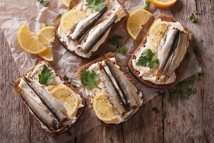 four slices of brown bread topped with two mackerel on a creamy dressing topped with fresh lemon wedges and herbs 