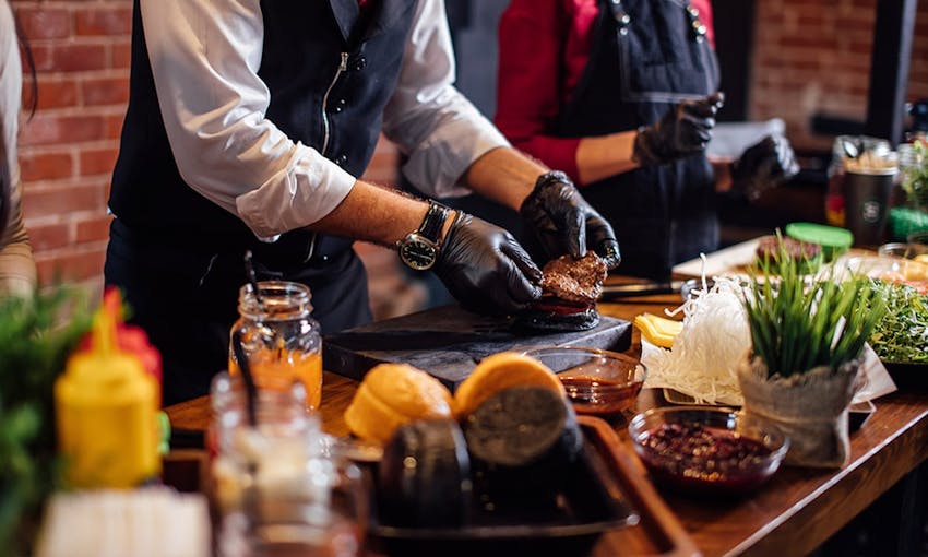  long wooden table with a range of plated foods set up for alive cooking demonstration where a man in a suit and black gloves places a joint of meat onto a chopping board 