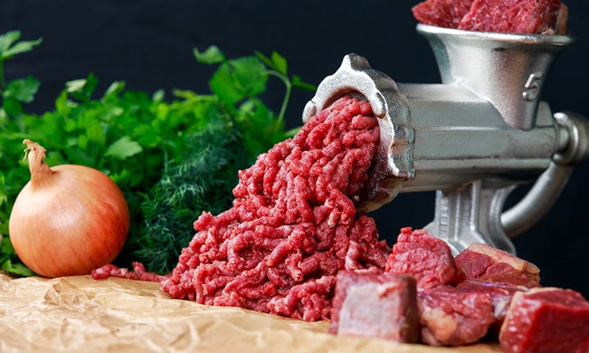 reddish meat being grinded in a silver meat grinder surrounded by fresh onion, chunks of red meat and fresh green herbs 