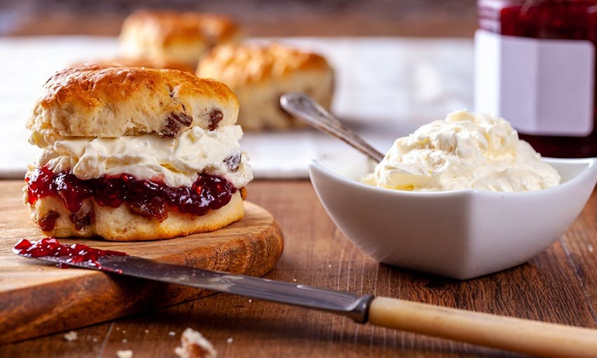 golden baked scone split in half and filled with a layer of sweet raspberry jam and a layer of thick cream on a wooden chopping on a wooden table next to a square bowl filled with cream 