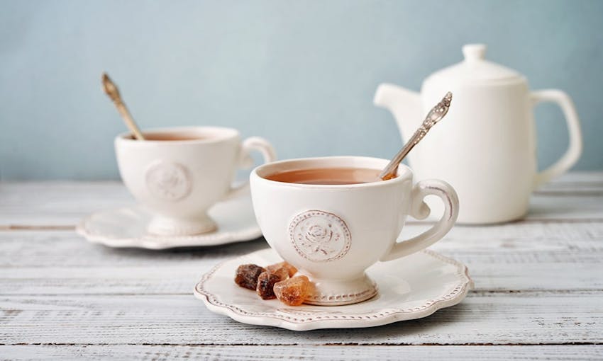 two teacups with a rose emblem on saucers filled with english breakfast tea with a brass spoon in the middle for stirring and a side of sugar cubes