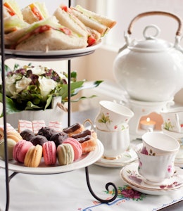 afternoon tea set up complete with a white chine teapot and teacup set on a white flowery tablecloth and a tiered plate stand with macarons, angel slices, eclairs and truffles on the bottom and mini sandwiches on top 