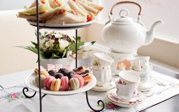 afternoon tea set up complete with a white chine teapot and teacup set on a white flowery tablecloth and a tiered plate stand with macarons, angel slices, eclairs and truffles on the bottom and mini sandwiches on top 