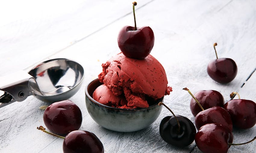 two scoops of cherry ice cream in a small grey bowl next to an ice cream scooper topped with brandy soaked cherries 