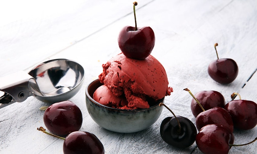 Cherries soaked in brandy make for a perfect dessert topping two scoops of cherry ice cream in a small grey bowl next to an ice cream scooper topped with brandy soaked cherries