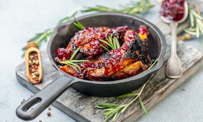 a small skillet with fried chicken wings glazed with a cherry reduction on top of a chopping board and decorated with fresh herbs salt and pepper 