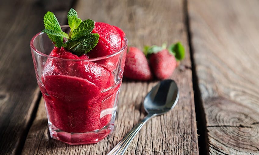 a small jar of bright cherry coloured sherbet with mint leaves and a silver teaspoon on top of a wooden table 