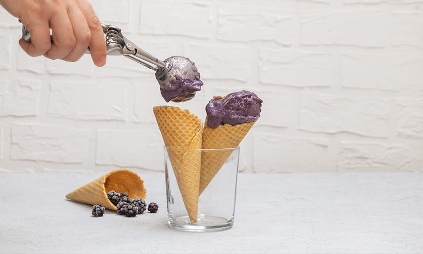 person spooning scoops of blackberry gelato onto two ice cream cones in a glass jar on white table next to an ice cream cone with frozen blackberries 