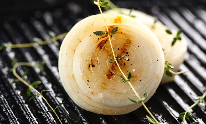 half of a white onion on top of a bbq grill next to two small shoots of herbs 