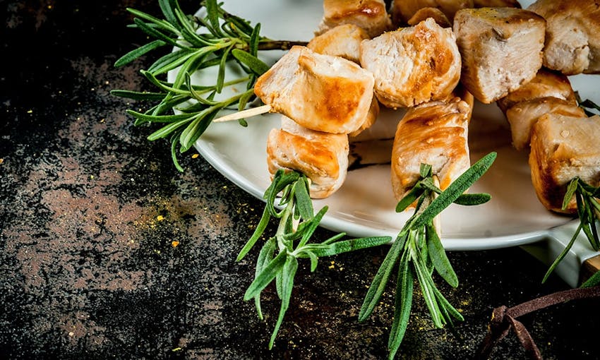 diced chicken breast cubes on rosemary skewers on a white plate on a dark wooden table top 