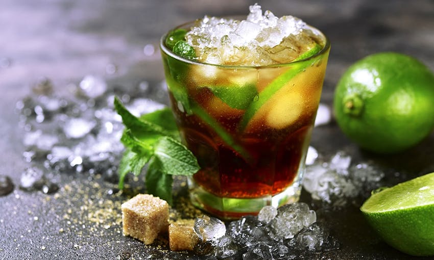 a small glass of ice tea with fresh mint topped with shaven ice surrounded by shaven ice, brown sugar cubes and fresh limes on a dark charcoal table top