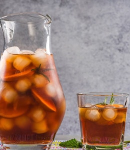 a two litre jug filled with ice cubes, fruit and peach iced tea and next to a small glass of peach iced tea with fresh sprig of mint 