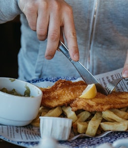 Fish & Chips man in grey jumper tucks in to a plate of fish and chips on british newspaper with a side of mushy peas topped with a wedge of lemon