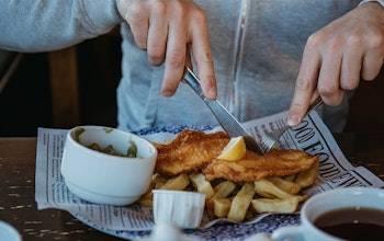 Fish & Chips man in grey jumper tucks in to a plate of fish and chips on british newspaper with a side of mushy peas topped with a wedge of lemon
