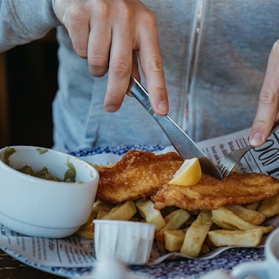 man in grey jumper tucks in to a plate of fish and chips on british newspaper with a side of mushy peas topped with a wedge of lemon 