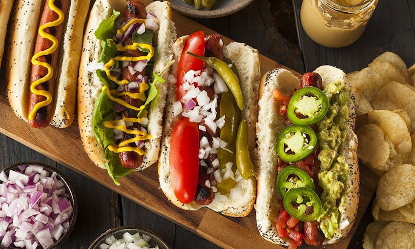 four hot dogs one topped with jalapenos and guacamole, one with gherkins and peppers, one with mustard and onions and the other with mustard next to crisps and a dish of onions 