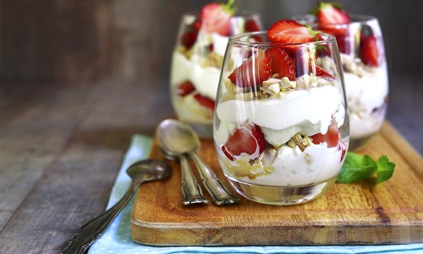 Three clear jars filled with eton mess made up of cream yoghurt strawberries and meringue on a wooden shopping board 