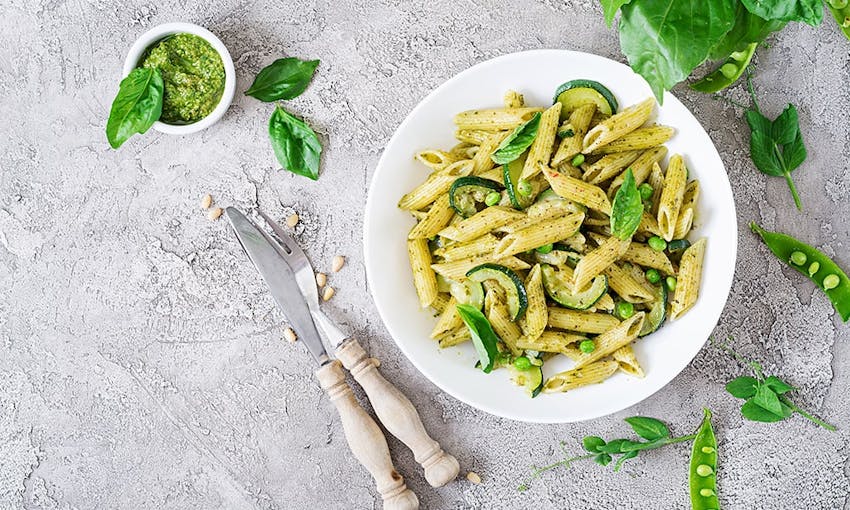 white bowl filled with pesto pasta with courgette, peas and topped with fresh herbs next to a dish of handmade pesto on a marble top table 