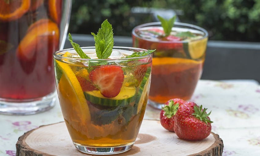two clear glasses of pimms filled with fresh fruits on a summer dining table next to a jug of pimms 