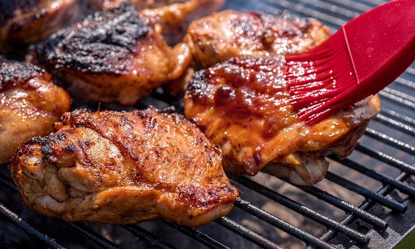 crispy chicken thighs on a bbq being basted with a bbq sauce with a red brush 