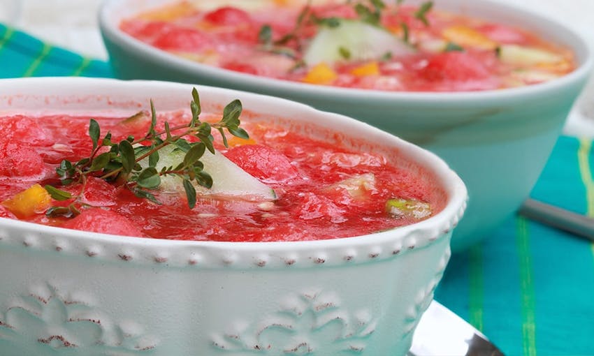 two china engraved bowls of cold summery watermelon soup topped with fresh herbs on a bright blue and green tablecloth 