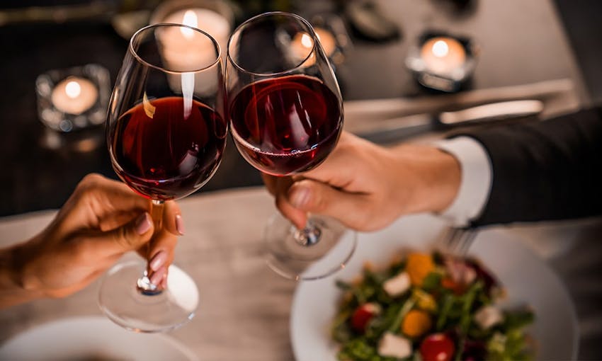 A man and a lady clinking two glasses of red wine over a candle lit restaurant meal 