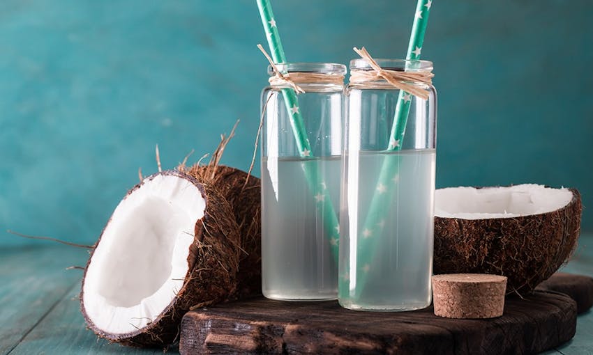 two coconuts halved next to a chopping board with two clear tumblers filled with coconut water with blue straws with a star pattern 