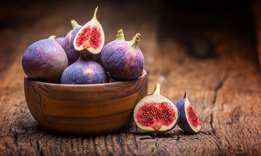 a wooden bowl with a pile of purple figs, some whole, some halved on top of a wooden table 