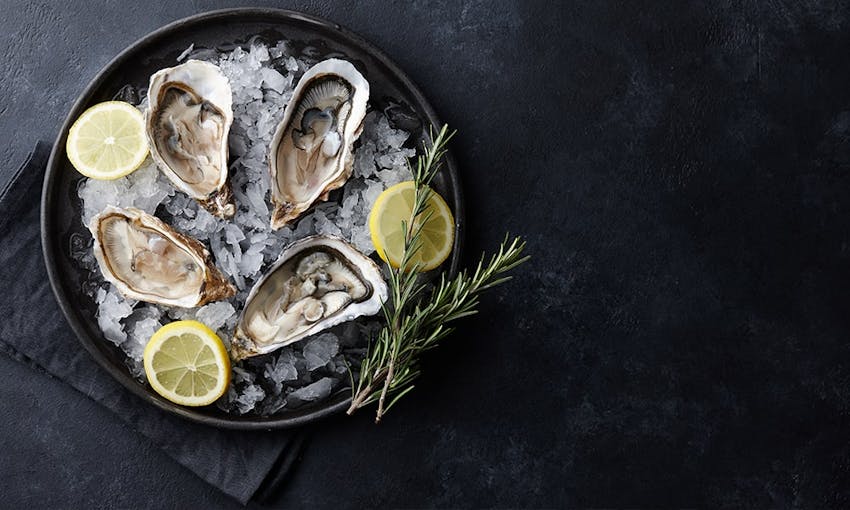 four oysters placed in a dark black bowl on top of shaven ice with two sprigs of rosemary and three wedges of lemon 