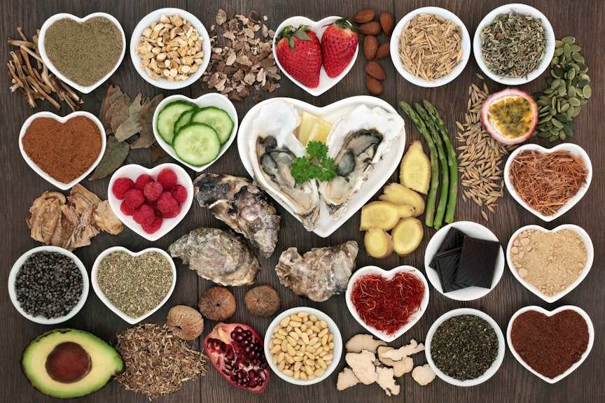 a birdseye view of different aphrodisiac foods, strawberries, asparagus, dark chocolate, cucumber, avocado and seeds in small circular and heart shaped dishes on a wooden table 