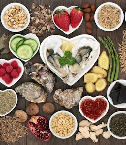 a birdseye view of different aphrodisiac foods, strawberries, asparagus, dark chocolate, cucumber, avocado and seeds in small circular and heart shaped dishes on a wooden table 