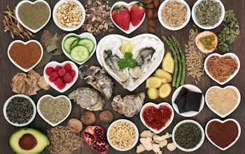 a birdseye view of different aphrodisiac foods, strawberries, asparagus, dark chocolate, cucumber, avocado and seeds in small circular and heart shaped dishes on a wooden table 