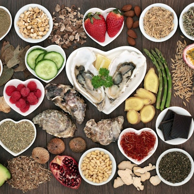a birdseye view of different aphrodisiac foods, strawberries, asparagus, dark chocolate, cucumber, avocado and seeds in small circular and heart shaped dishes on a wooden table 
