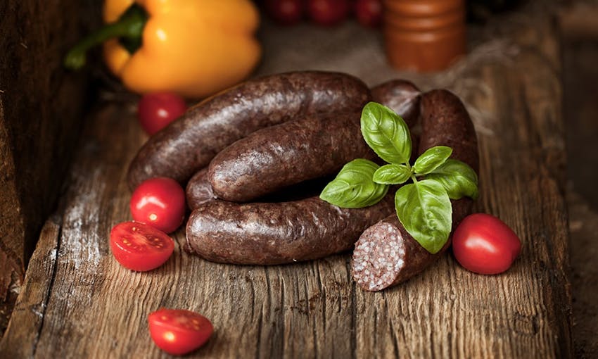 four black pudding sausages on an oak wooden chopping board with half diced cherry tomatoes and a yellow pepper in the background  