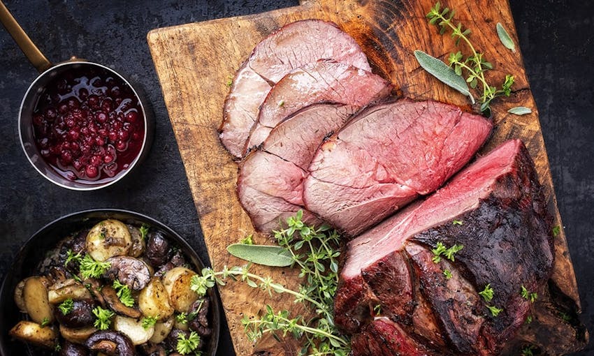 a roasted joint of venison on a wooden chopping board with a small saucepan of cranberry sauce and a side dish of roast potatoes and mushrooms 