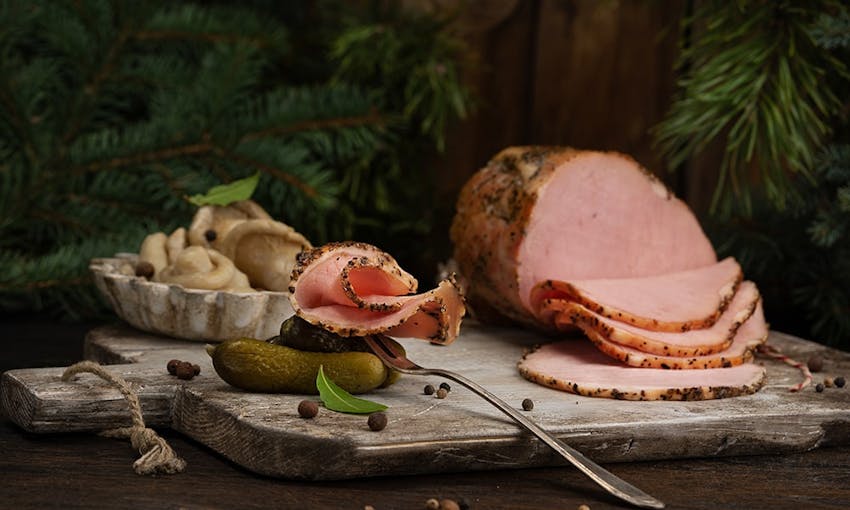 whole christmas ham partially sliced on a grey wooden chopping board with mushrooms in a side dish along with gherkins and sprinkled peppercorns 