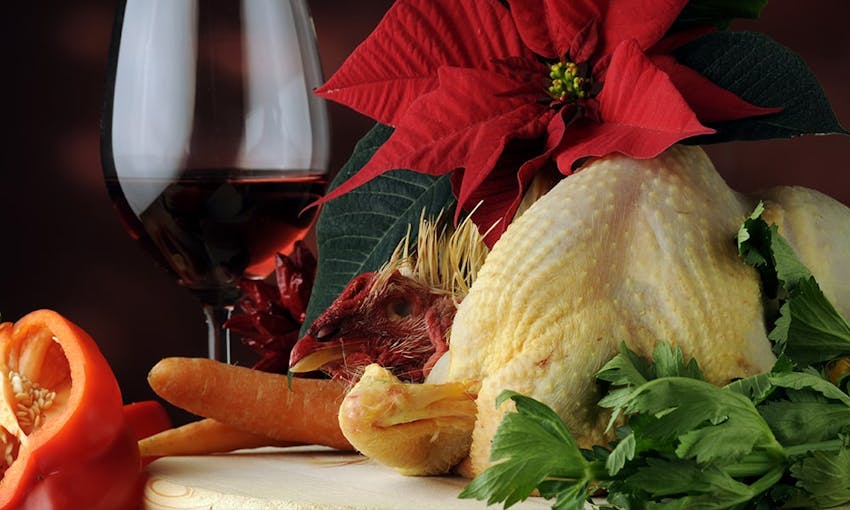 a capon on a white chopping board surrounded by fresh herbs peppers carrots and a christmas wreath red flower and a glass of red wine 
