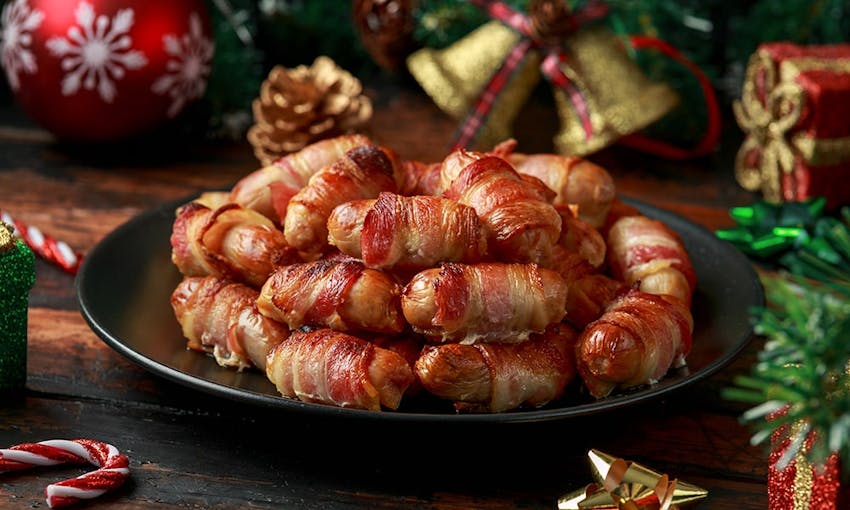 small cocktail sausages wrapped in cripsy bacon stacked high on a black plate surrounded by christmas table decorations 